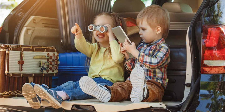 Two toddlers in an open car boot playing with toy binoculars and a tablet before a long drive