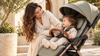Mother smiling at her toddler seated in a premium sage green pushchair with a safety harness, surrounded by warm natural sunlight and greenery.