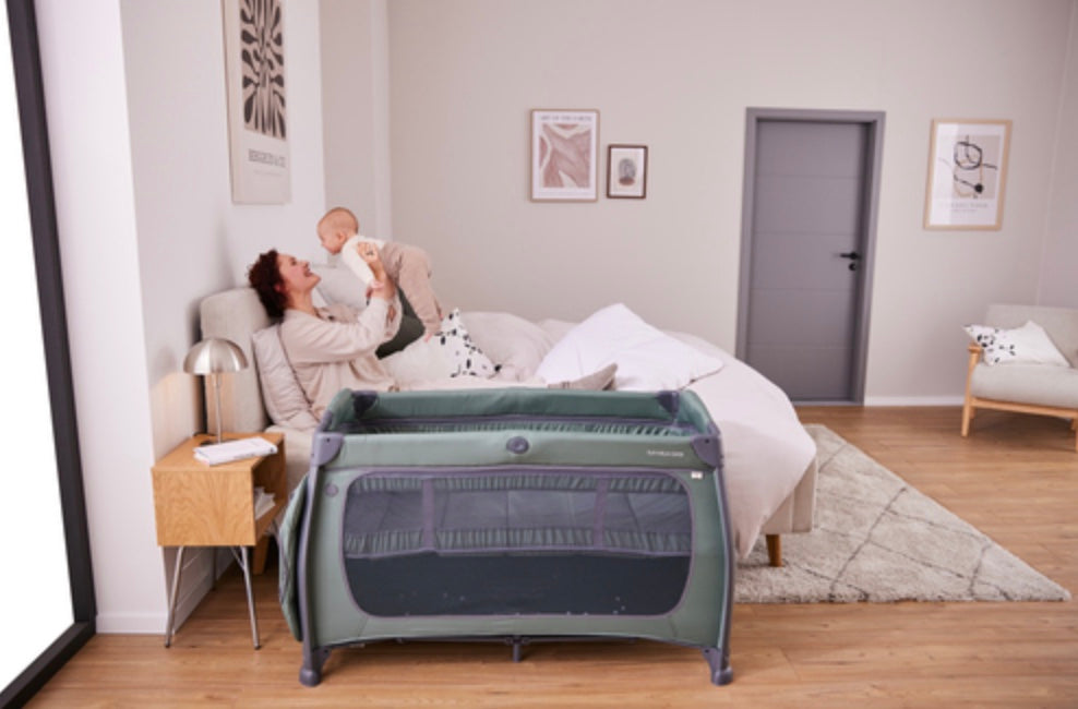 Hauck Play N Relax Center travel cot in Dark Green set up next to the bed while a mother lifts her baby